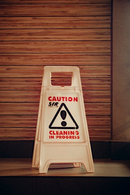 A white, portable 'Cleaning in Progress' caution sign with a red and black warning symbol, placed on a wooden floor next to a wall with horizontal wooden paneling. The scene is illuminated with natura