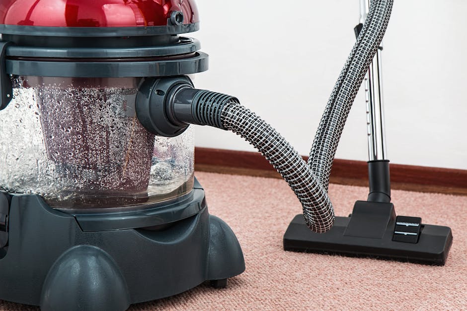 A professional cleaner wearing a white protective suit, face mask, and blue gloves is performing deep cleaning and sanitisation of a beige carpet inside a well-lit living room. The cleaner is using a
