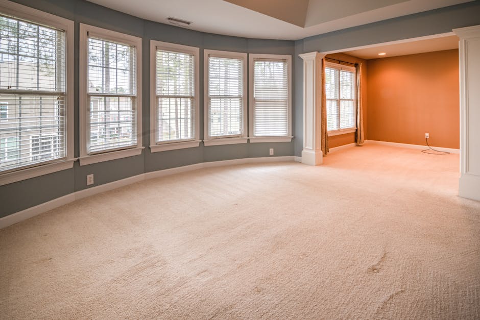 A person in orange work overalls vacuuming a light-colored tiled floor in a modern living room, with a yellow vacuum cleaner featuring a black handle and hose. The room includes a dark gray fabric sof